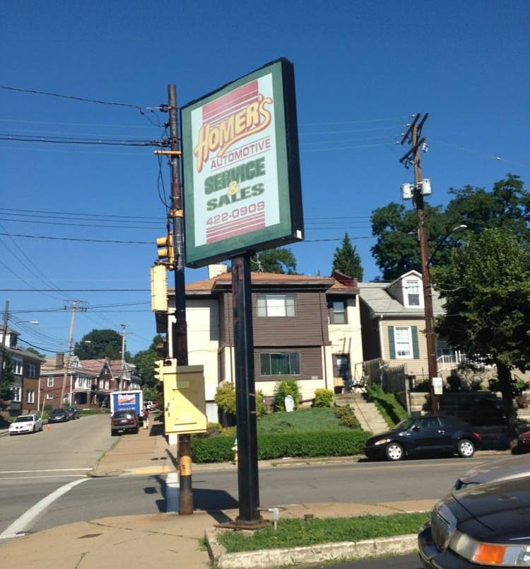 Close view of the green-framed Homer&rsquo;s Automotive Service & Sales sign, showing the phone number 422-0909, on its pole beside the street.